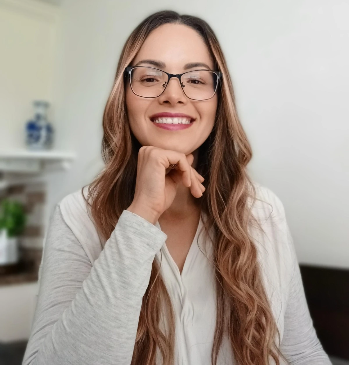 A smiling woman with long, wavy brown hair and glasses, resting her chin on her hand against a soft-focus indoor background.